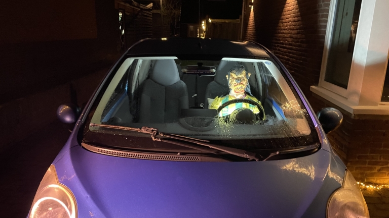 Een blauwe auto geparkeerd in een smalle steeg, met een halloweenmasker van een monster op de bestuurdersstoel zichtbaar door de voorruit, 's nachts gefotografeerd met lichte regen op de ramen.
