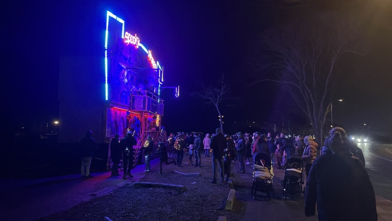 Een groep mensen verzamelt zich 's avonds buiten voor een verlicht attractiegebouw met felle neonkleuren, terwijl sommigen kinderwagens bij zich hebben.