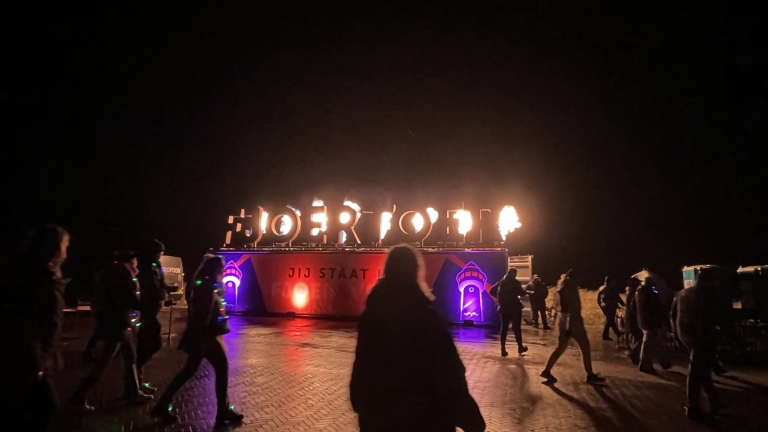 Een nachtelijk tafereel met mensen die voor een verlichte installatie staan, waarop brandende letters zichtbaar zijn.