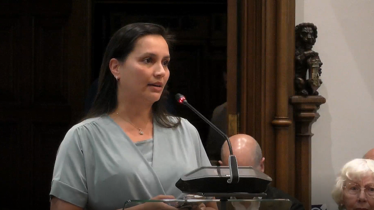 Een vrouw spreekt voor een microfoon in een formele setting, met een houten pilaar en een decoratief beeldhouwwerk op de achtergrond. Er zit een oudere vrouw met wit haar rechts op de voorgrond.