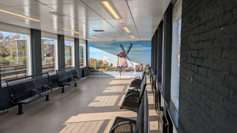 Een wachtruimte op een treinstation met zwarte banken en brede ramen waardoor buiten de spoorlijnen zichtbaar zijn. Aan de muur hangt een groot schilderij van een traditionele Nederlandse windmolen bij een waterkant.