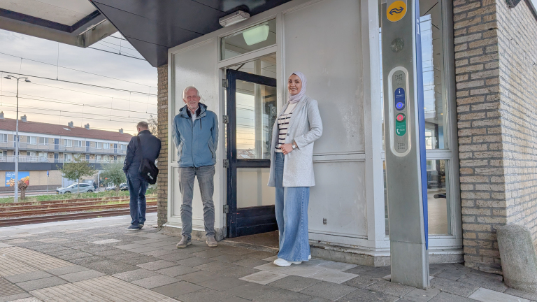 Een man en vrouw staan ​​lachend voor een station met NS-logo, met een hulppaal en treinspoor op de achtergrond.