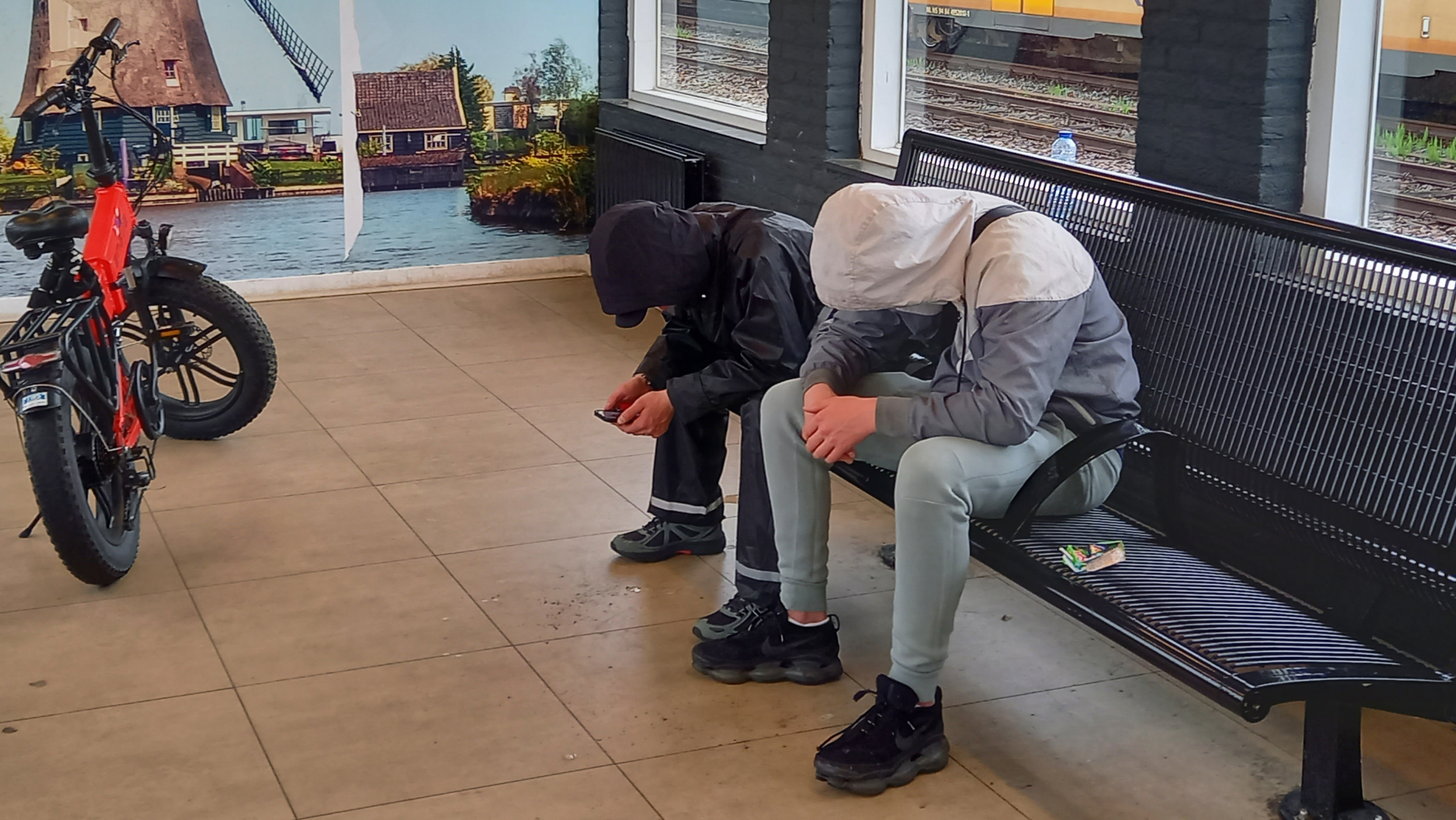 Twee personen met capuchons zitten in een wachtruimte op een bankje, dicht bij een rode fiets. Op de achtergrond is een afbeelding van een molen te zien en een voorbijrijdende gele trein buiten het raam.