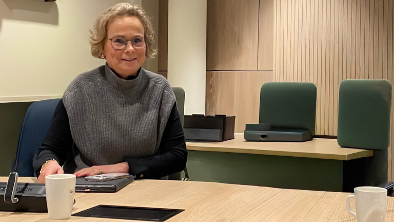 Een vrouw met bril zit aan een vergadertafel in een moderne kantoorruimte, met houten panelen en groene stoelen op de achtergrond.