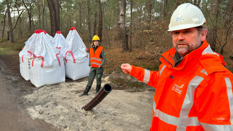 Twee mensen bij grote zakken grond in een bosachtige omgeving, gekleed in oranje veiligheidsjassen en helmen, met een pijp die uit de grond steekt.