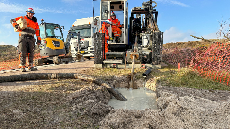 Bouwvakker in oranje veiligheidskleding draagt houten blokken naast een grijze graafmachine en ander bouwmaterieel op een bouwplaats met een uitgegraven sleuf gevuld met modderig water.