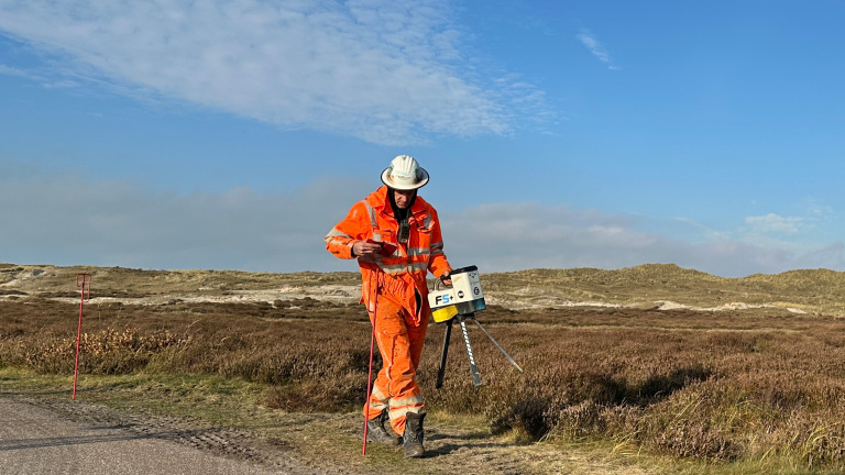 Een persoon in een oranje veiligheidsuitrusting, inclusief helm, voert landmeetwerkzaamheden uit met een meetapparaat in een open, heuvelachtige gebied onder een heldere blauwe hemel.
