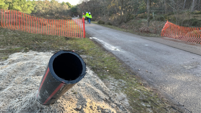 Een plastic buis ligt in een hoop zand naast een weg met rode veiligheidshekken; in de verte lopen mensen in veiligheidskleding.