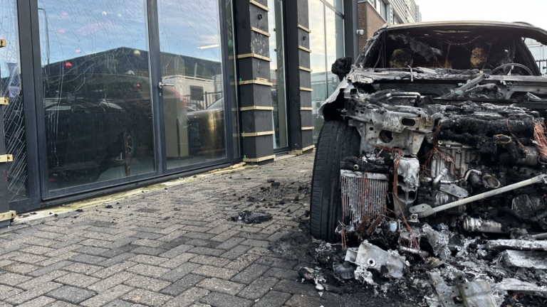 Een uitgebrande auto naast een gebouw met gebarsten ramen, omgeven door verbrandde puinresten op de grond.