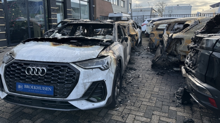 Uitgebrande auto's op een parkeerterrein voor een autobedrijf, met zwaar beschadigde carrosserieën en geblakerde oppervlakken.