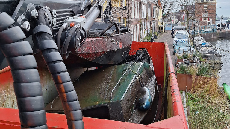 Een kraan tilt een klein, vuil schip uit het water en plaatst het in een rode bak in een smalle straat naast een kanaal met geparkeerde auto's en boten, omringd door bakstenen gebouwen.