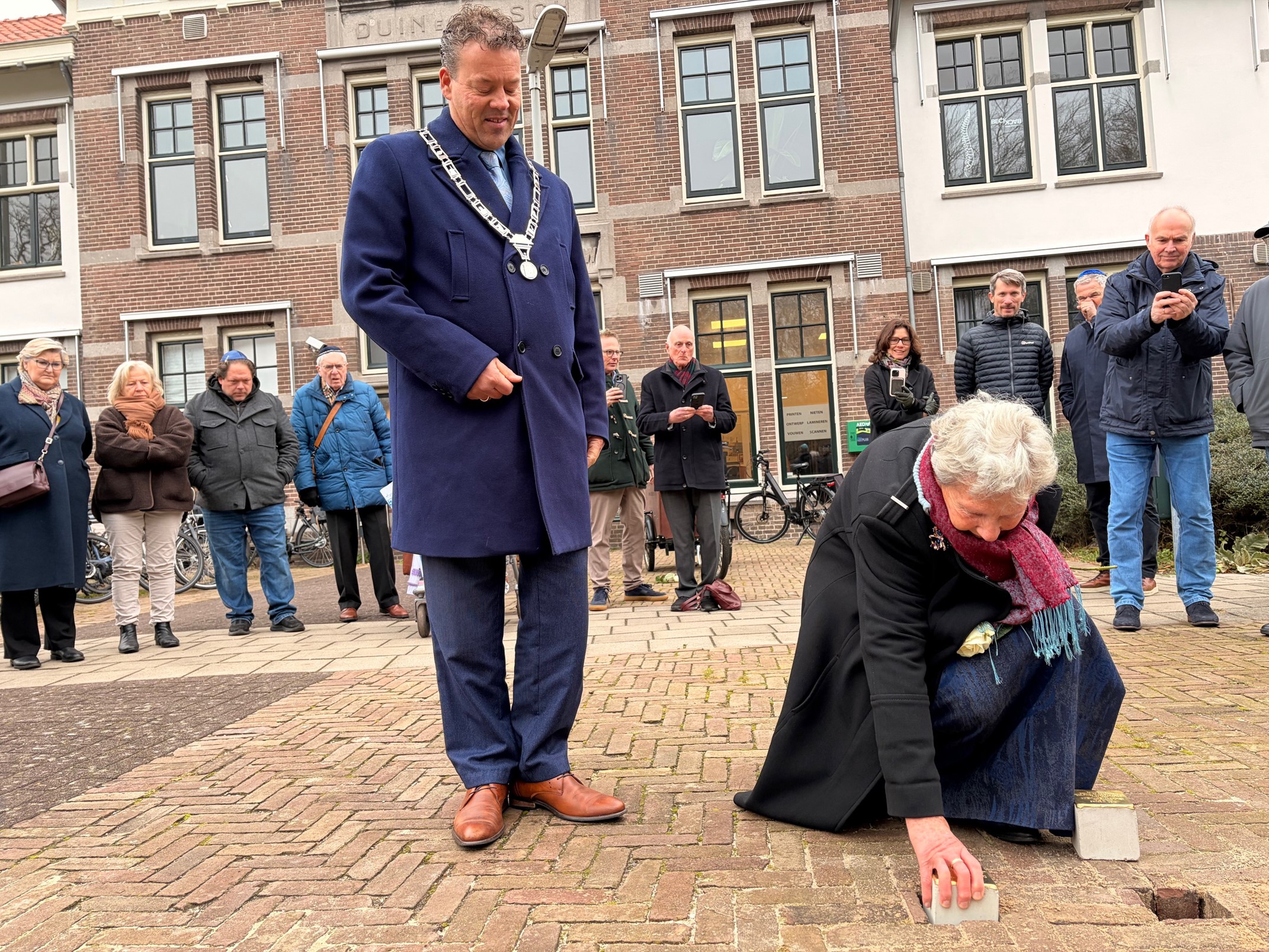 Een vrouw buigt zich voorover om een herdenkingssteen te plaatsen op een plein, terwijl een man in een blauwe jas toekijkt en een groep mensen op de achtergrond toekijkt.