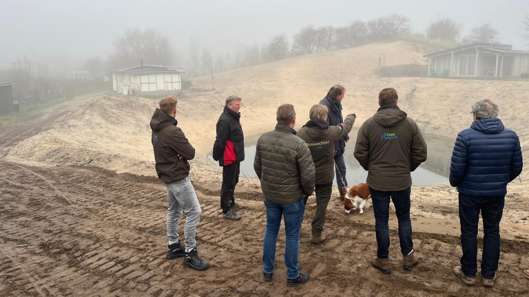 Een groep mensen staat in mistige omstandigheden rond een kunstmatige vijver in een zandachtig landschap, met een hond bij hen, terwijl iemand aanwijzingen geeft.