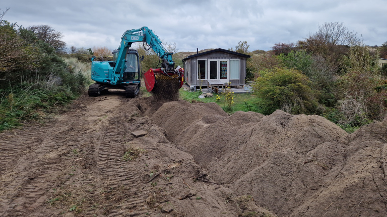 Een blauwe graafmachine met een rode graafbak verplaatst zand in de buurt van een houten huisje omgeven door struiken en bomen.