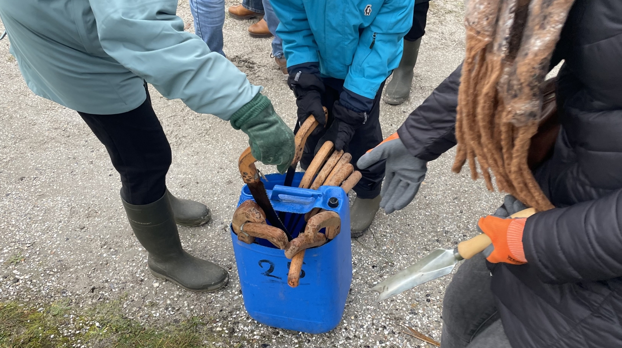 Mensen in buitenkleding pakken houten handgereedschap uit een blauwe container met het getal '2' erop geschreven.