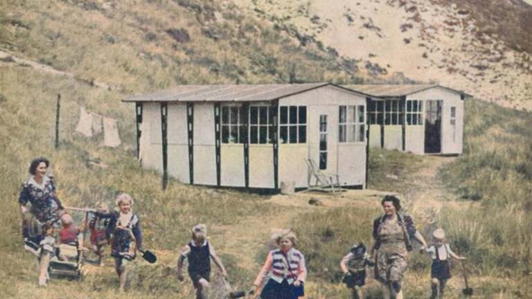 Een groep vrouwen en kinderen loopt in een grasrijk gebied voor een eenvoudige witgeschilderde barak met ramen. Op de achtergrond hangen kleren aan een waslijn naast de barak.