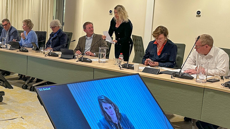 Mensen zitten rond een conferentietafel tijdens een vergadering, met een scherm dat een spreker toont en "03 Debat" aangeeft.