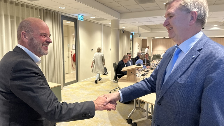 Twee mannen in pakken schudden elkaar de hand in een vergaderruimte met mensen zittend aan een tafel op de achtergrond.