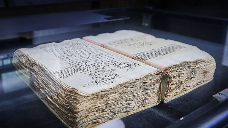 Een oud, dik boek met handgeschreven tekst en versierde handtekeningen op de pagina's, vertoond onder een glazen dekking.