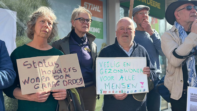 Een groep mensen met protestborden bij een demonstratie. Eén bord zegt 'Statushouders wonen ook graag droog' en een ander bord zegt 'Veilig en gezond wonen voor alle mensen'.