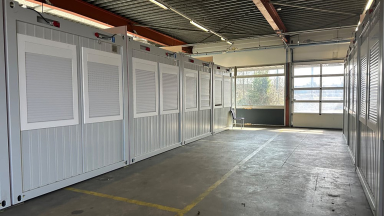 Een lege binnenruimte met meerdere witte containers langs de zijwand, betonnen vloer en grote ramen aan het einde van de ruimte.