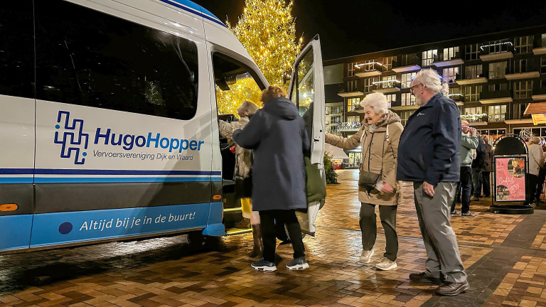 Mensen stappen 's avonds in een HugoHopper busje naast een verlichte kerstboom, op een plein met verlichte gebouwen.