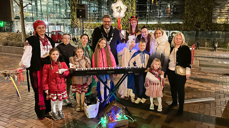 Een groep vrouwen en kinderen, sommigen gekleed in traditionele kleding, poseren 's avonds buiten rond een keyboard en een miniatuur kersttafereel, met een gebouw op de achtergrond.