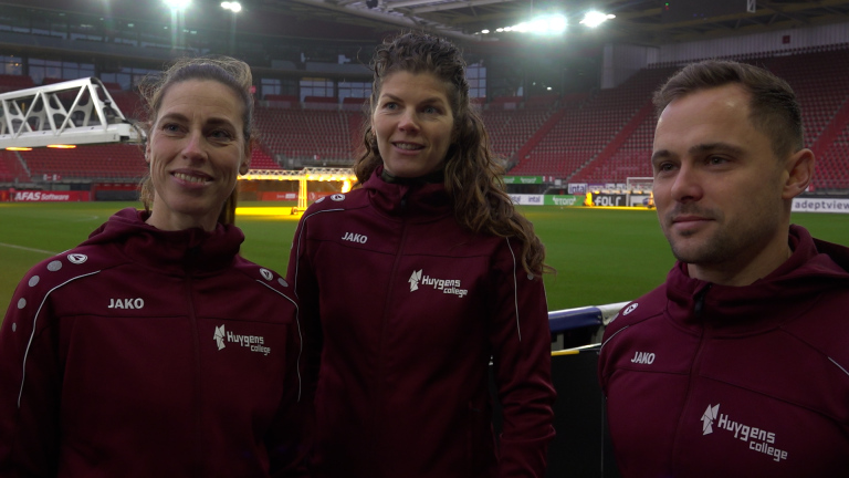 Drie mensen in trainingsjacks met 'Huygens College' logo staan in een stadion met lege tribunes op de achtergrond.