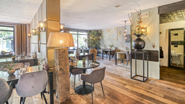 Een sfeervol ingerichte eetkamer met houten vloeren, moderne tafels en stoelen, grote vazen met bloemen en planten, en glazen deuren aan de zijkant die uitkijken op de tuin.