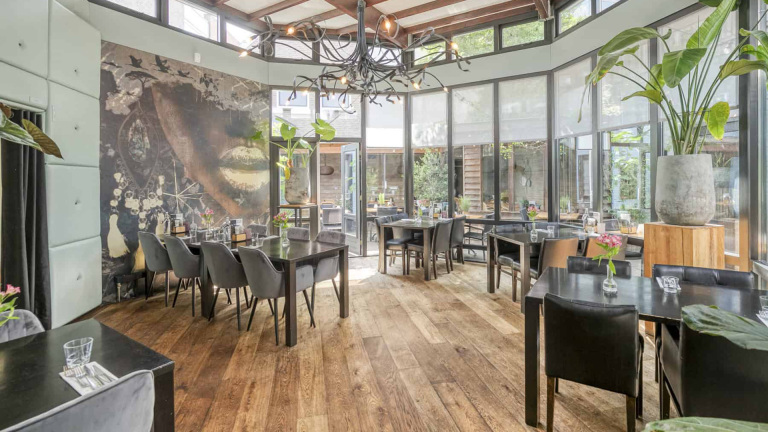 Een lichte, moderne eetruimte met houten vloeren, decoratieve planten, en glazen ramen, ingericht met zwarte tafels en stoelen omgeven door een grote muurschildering en een opvallende zwarte kroonluchter aan het plafond.