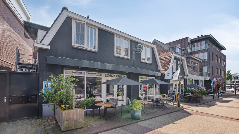Een zwart geschilderd café met een klein terras met parasols en tafels met planten aan een rustige straat.