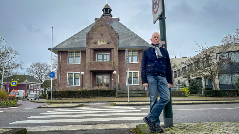 Een man leunt tegen een verkeersbord op een trottoir voor een historisch gebouw met het jaartal 1927 op de gevel.