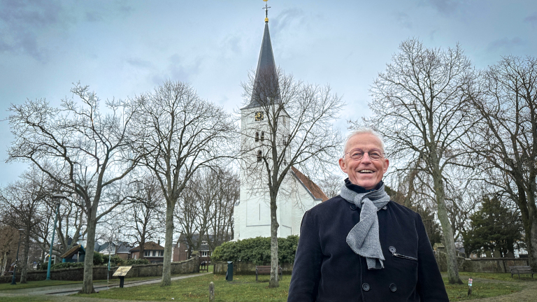 Een man met grijze haren en een sjaal staat glimlachend voor een kerk met een hoge toren, omringd door bomen zonder bladeren.