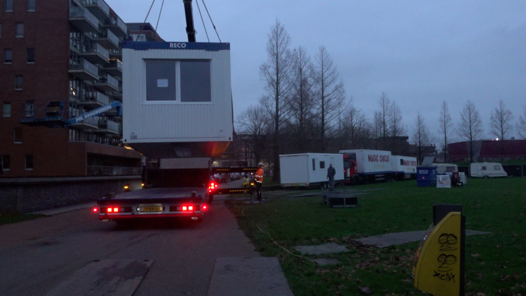 Een grote container van RECO wordt met een hijskraan verplaatst in een stedelijke omgeving, omringd door vrachtwagens van Magic Circus en enkele mensen op de grond.