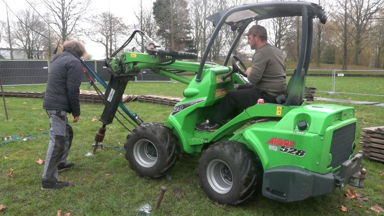 Een man bedient een Avant 528 mini loader op een grasveld, terwijl een andere man naast het voertuig staat en lijkt te assisteren bij het boren van een paal in de grond. Op de achtergrond zijn hekken en opgestapelde planken zichtbaar.