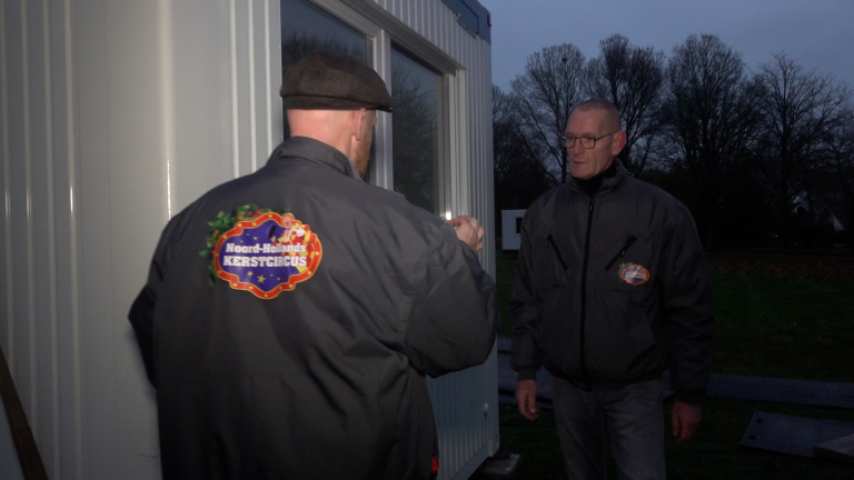 Twee mannen staan 's avonds buiten bij een gebouw, gekleed in jassen met een logo van 'Noord-Hollands Kerstcircus'.