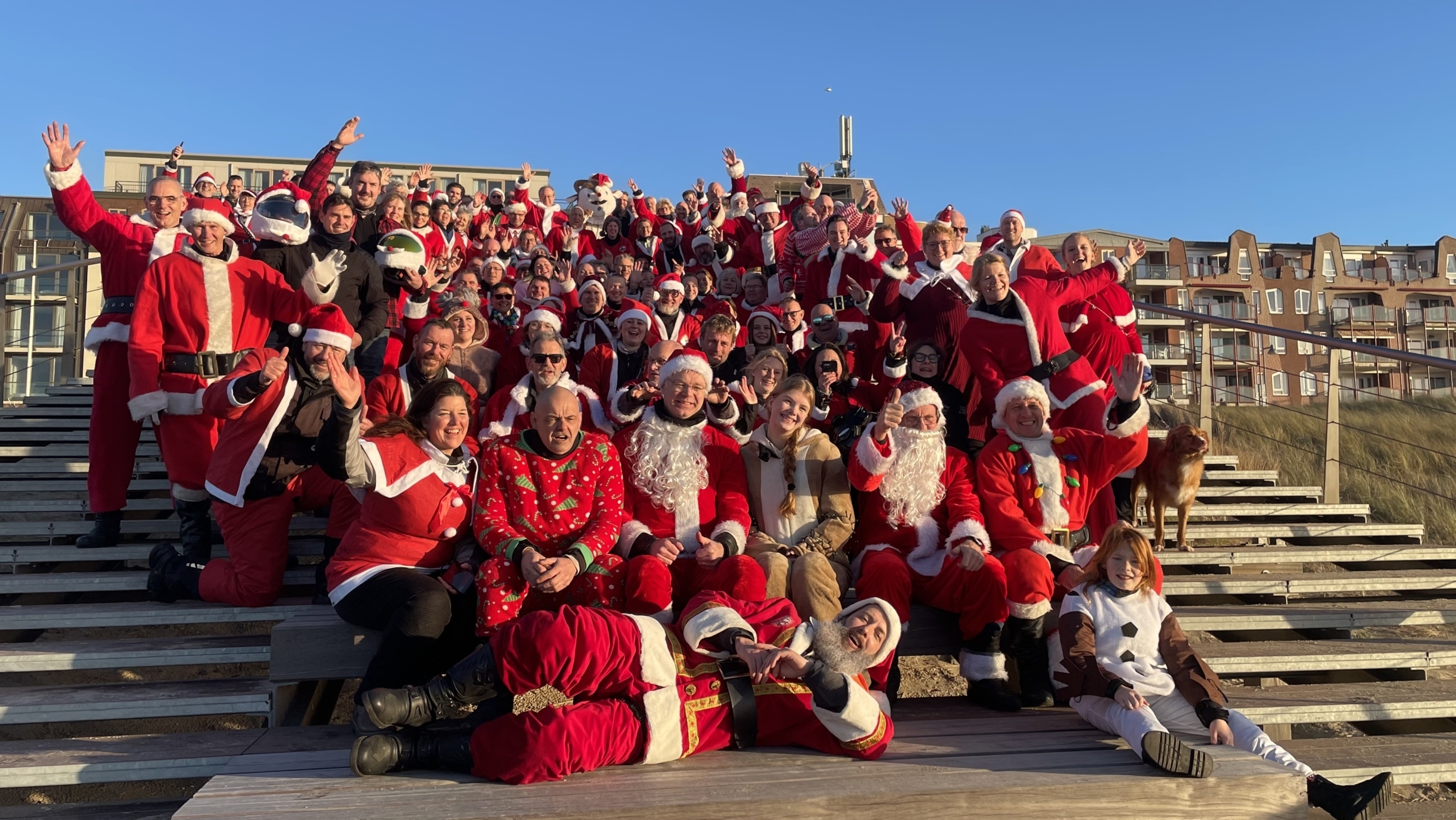 Grote groep mensen gekleed in kerstmannenpakken poseren op een houten trap, met een heldere blauwe lucht en appartementen op de achtergrond. Een kind en een hond zitten vooraan.