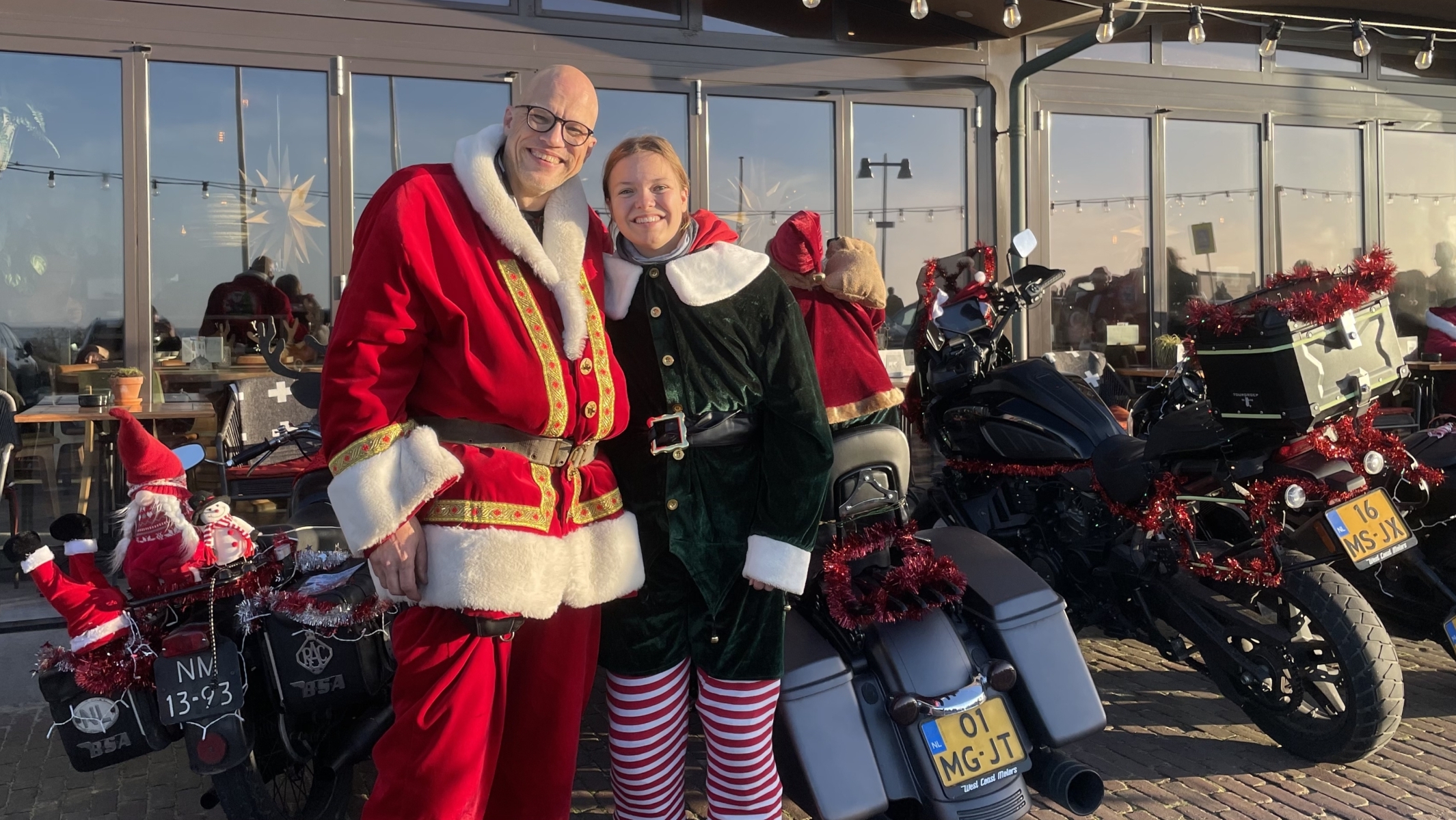 Twee personen gekleed als kerstman en elf poseren voor een café met versierde motoren op de achtergrond.