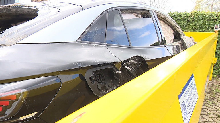 Beschadigde elektrische auto in een gele container, zichtbaar laadpoort, kapotte ramen en verwoest interieur.