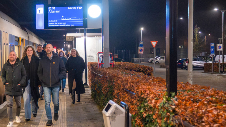 Een groep mensen loopt 's nachts op een perron van een treinstation met een informatiebord dat de bestemming Alkmaar aangeeft.