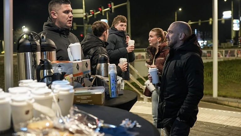 Een groep mensen in warme jassen staat 's nachts bij een tafel met koffie en snacks bij een kruispunt met verkeerslichten.