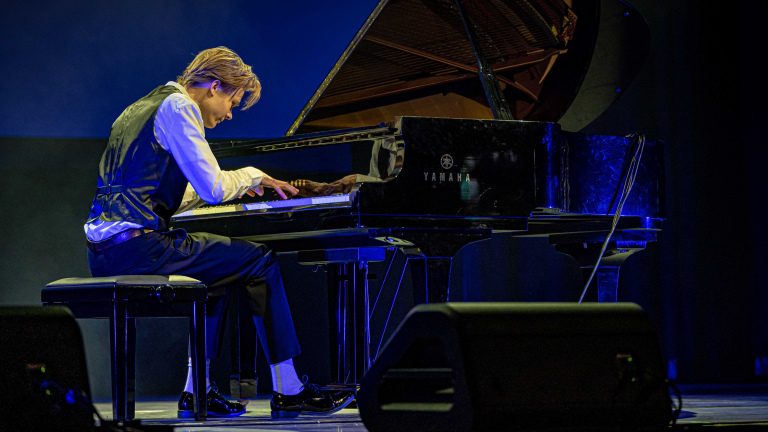 Een man in een vest en witte blouse speelt piano op een podium, onder blauwe belichting.
