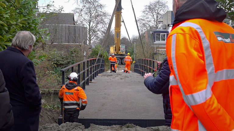 Bouwvakkers in fluorescerende oranje jassen werken aan de installatie van een brug met behulp van een grote kraan, terwijl omstanders toekijken.