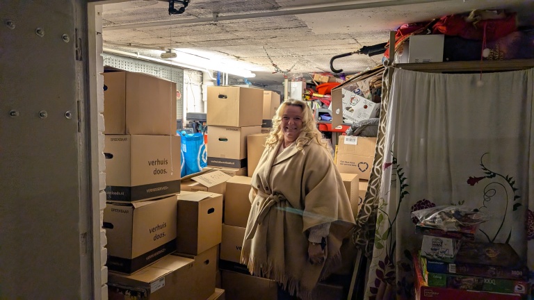 Vrouw met een beige mantel staat in een ruimte vol met opgestapelde verhuisdozen en speldozen.