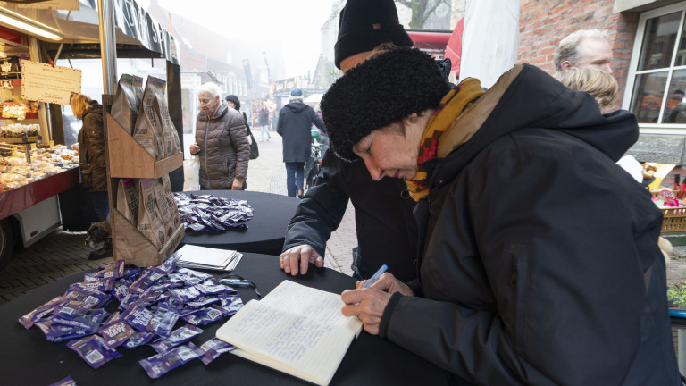 Vrouw schrijft in een notitieboekje bij een marktkraam met pakjes en snacks op tafel, terwijl andere marktkramen zichtbaar zijn op de achtergrond.