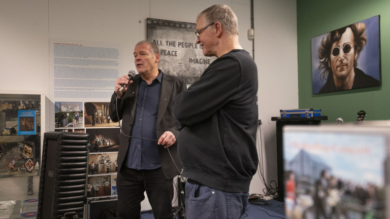 Twee mannen praten in een kamer met posters en een schilderij van een man met zonnebril aan de muur.