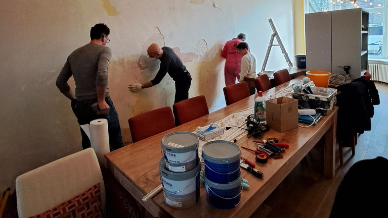 Mensen werken aan een muur in een ruimte met stoelen, tafels vol gereedschap en potten verf.