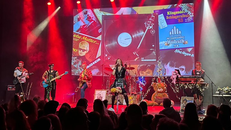 Een band treedt op op een podium met een achtergrond vol kleurrijke vintage afbeeldingen, terwijl rode lichten de sfeer bepalen.