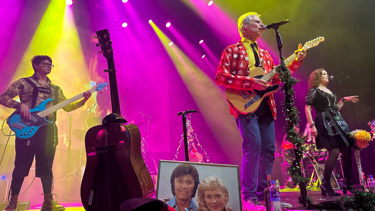Muzikanten spelen op een podium met gekleurde lichten, een gitarist in een kersttrui en een zangeres in een traditionele jurk.