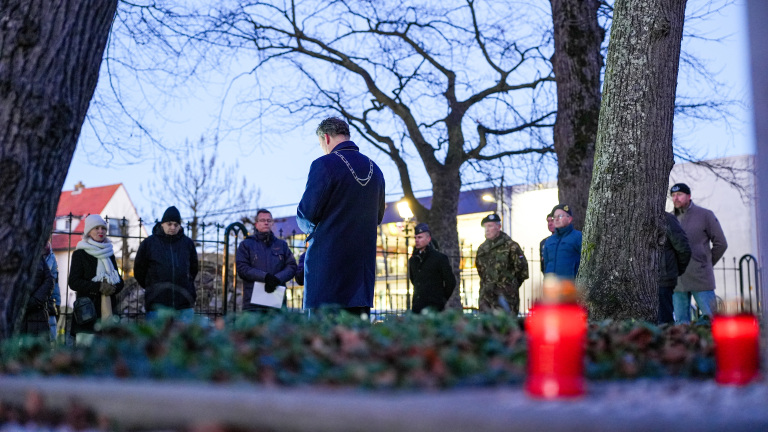 Mensen staan bij een herdenking in een park, met een man in een blauwe jas en fakkels op de voorgrond.